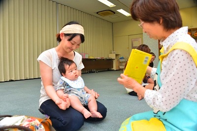 お母さんの膝の上でスタッフの読み聞かせ絵本を見る赤ちゃんの画像