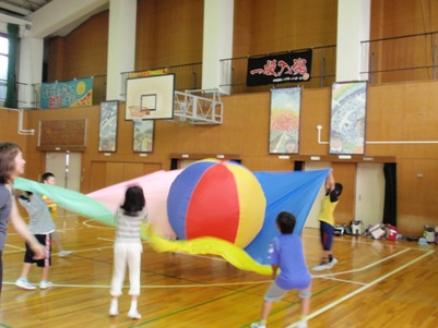 体育館で巨大ボールをシートで飛ばす遊びをする児童たち