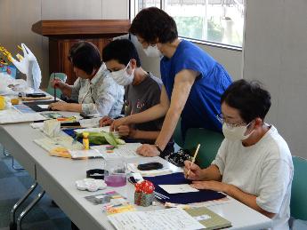 横に並んで座っている受講者の間に先生が入って絵手紙の指導をしている