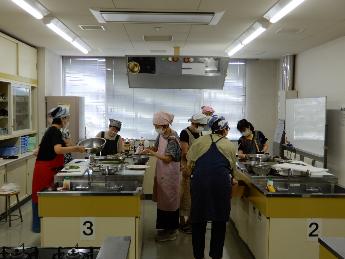 料理教室でグループに分かれて作業台で食材を切ったりコンロを使って調理している