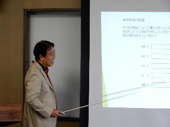 先生がスクリーンに映し出された資料をさし棒を使って説明しているところ